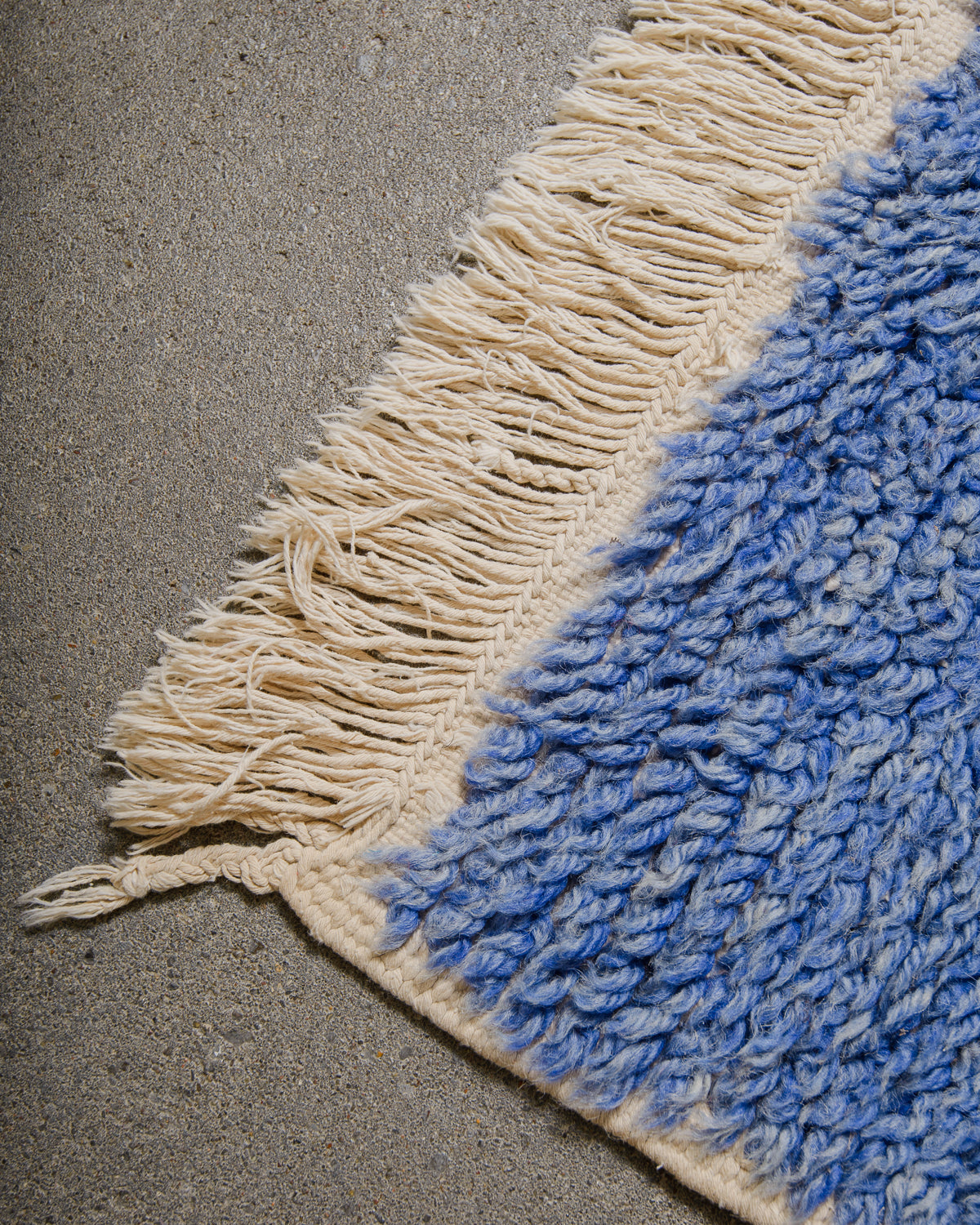 Close-up of a blue and cream textured rug corner with tassels on a concrete floor.